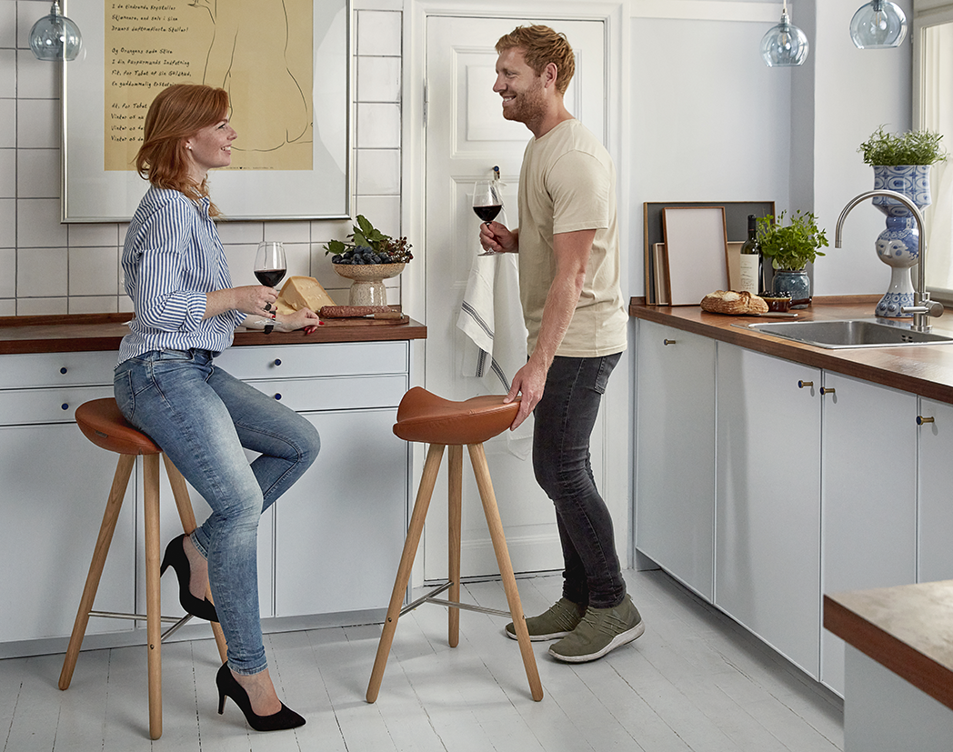 Kvinde og mand opholder sig i et køkken. Kvinden sidder på en barstol, CCL High, betrukket med cognacsfarvet skind. Manden er ved at sætte sig på sin barstol hvor de sammen kan nyde at glas rødvin.